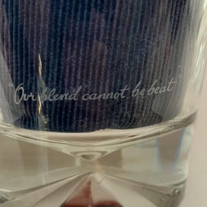 Pu&ograve; includere: Un bicchiere trasparente con una base decorativa e la scritta incisa "Our blend cannot be beat". Il bicchiere contiene un materiale blu scuro strutturato. Il bicchiere &egrave; su una superficie bianca.