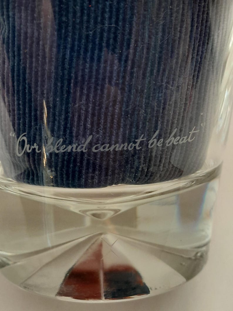 Pu&ograve; includere: Un bicchiere trasparente con una base decorativa e la scritta incisa "Our blend cannot be beat". Il bicchiere contiene un materiale blu scuro strutturato. Il bicchiere &egrave; su una superficie bianca.