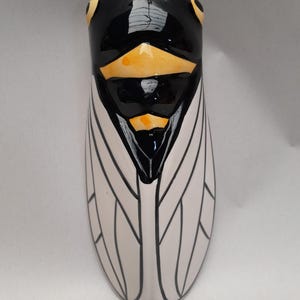 Op de afbeelding: Een keramieken cicadebeeldje met een zwarte kop en lichaam, gele accenten en witte vleugels met zwarte aderdetails. Het insectvormige kunstwerk is waarschijnlijk een decoratief item.
