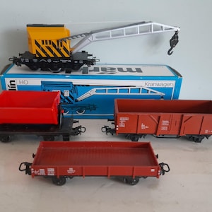 Puede incluir: Un conjunto de vagones de tren modelo, que incluye un vagón grúa amarillo y negro, un vagón tolva rojo y dos vagones de carga rojos. El conjunto se muestra con una caja azul que dice "Kranwagen".