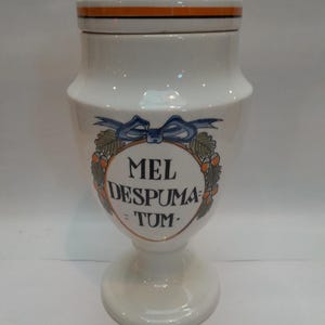 Op de afbeelding: Witte keramische pot met deksel, versierd met een blauw lint en oranje en bruine eikels. De pot heeft de tekst "MEL DESPUMA - TUM" aan de voorkant.