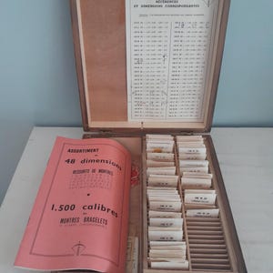 Può includere: Scatola di legno aperta contenente parti di riparazione di orologi. La scatola ha un coperchio incernierato con una tabella di riferimento. All'interno, ci sono numerose piccole carte e un libretto rosa con testo francese. Il libretto recita "ASSORTIMENT de 48 dimensions".