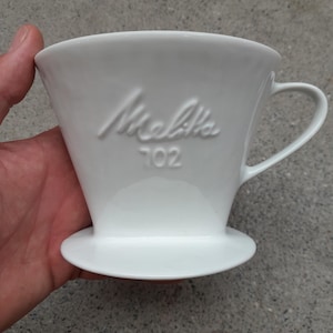 May include: A white ceramic coffee pour-over dripper with a handle and a wide circular base. The front of the dripper features the embossed text Melitta and the number 702. The design includes a wide top opening that tapers down to the base.