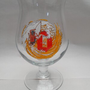 May include: A clear glass beer glass with a red and yellow design of a surfer riding a wave. The design includes the text "Sur la Mer".