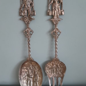 May include: A set of two silver-plated serving utensils with ornate handles. The handles feature figures of a man and a woman carrying buckets. The utensils are a spoon and a fork.