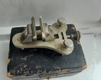 May include: A silver-toned watchmaker's vise with three knobs, resting on a black leather-covered box.