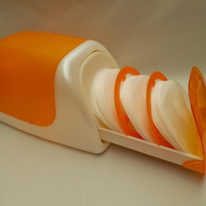 Op de afbeelding: Een wit en oranje kunststof dispenser met een stapel ronde, witte papieren filters. Het apparaat heeft een gladde, gebogen behuizing met een uitschuifbare lade die de ronde filters rechtop houdt, afgedekt met een doorzichtig oranje deksel.