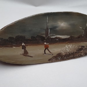 Op de afbeelding: Een handgeschilderd houten plaquette met een winterscène met twee figuren die schaatsen voor een kerktoren. De lucht is donkergrijs met een witte maan.