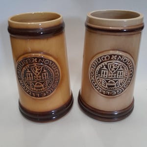 Puede incluir: Dos jarras de cerveza de cerámica con un esmalte marrón y beige. Cada jarra presenta un emblema circular con un escudo y el texto "SIGILLUM NOVUM". Las jarras tienen forma cilíndrica con la parte superior ligeramente ensanchada.