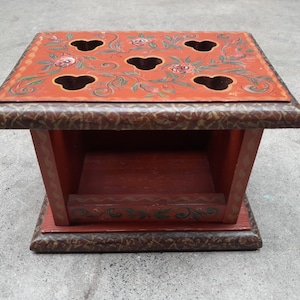 Puede incluir: Una caja decorativa cuadrada de madera roja y marrón con seis agujeros en forma de corazón en la parte superior. La parte superior está pintada con diseños florales en rosa, verde y blanco. Los lados y la base también están pintados con patrones decorativos.