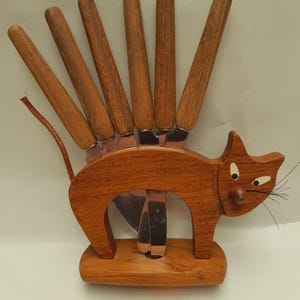 Op de afbeelding: Een houten messenset met een katvormige houder. De houder is gemaakt van bruin hout en heeft een katten silhouet met een staart en snorharen. Zes messen met houten handvatten worden in de houder getoond.