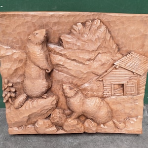 以下が含まれることがあります： 手彫りの木製レリーフ彫刻。山岳風景の中に2匹のマーモットが描かれています。1匹のマーモットは直立し、もう1匹は四つん這いになっています。小さな小屋と岩場も木に彫られています。彫刻は暖かく自然な茶色です。