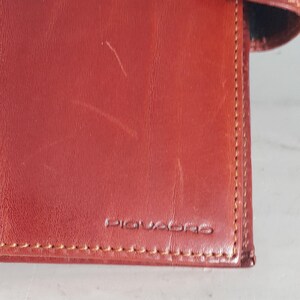 May include: Close-up of a reddish-brown leather wallet with the brand name "PIQUADRO" embossed on the front. The wallet has visible stitching and a slightly worn appearance.
