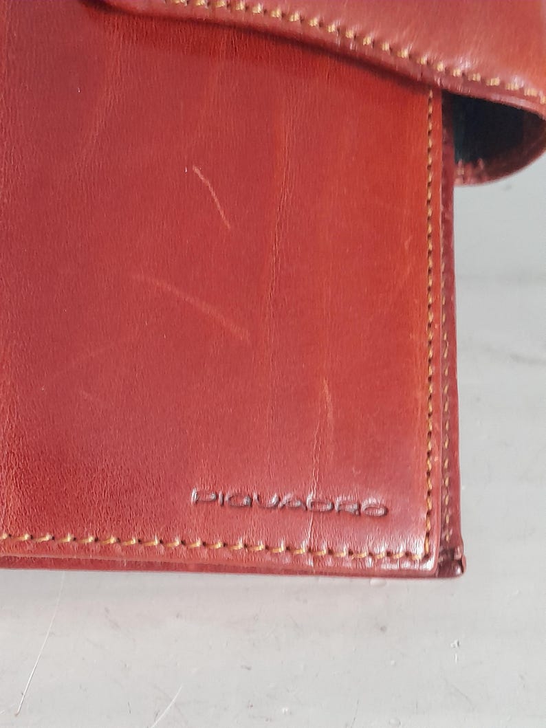 May include: Close-up of a reddish-brown leather wallet with the brand name "PIQUADRO" embossed on the front. The wallet has visible stitching and a slightly worn appearance.