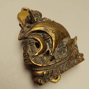 May include: A detailed, antique brass helmet-shaped door knocker. The door knocker features a knight's helmet design with intricate detailing, including a visor, crest, and decorative elements. The brass has a weathered appearance.