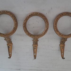 Op de afbeelding: Drie gouden metalen haken met sierlijke, ronde frames. Elke haak heeft een decoratief, gerold ontwerp.