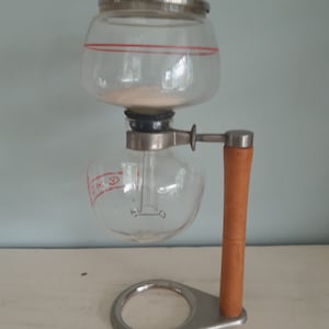 Pode incluir: Uma cafeteira de vidro com uma pega de madeira e uma base de aço inoxidável. A cafeteira tem uma jarra de vidro e um filtro de vidro. A cafeteira está sobre uma superfície branca.