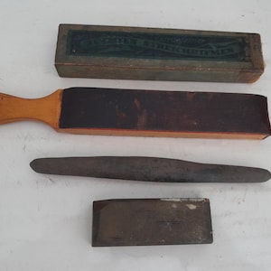 May include: An antique shaving kit with a wooden handle strop, a sharpening stone, a metal honing rod, and a box with text. The strop has a dark leather surface. The kit is likely from the early 20th century.