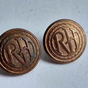 Peut inclure: Deux boutons anciens en bronze avec les lettres "RH" dans un motif circulaire. Les boutons présentent une surface texturée et une esthétique vintage. Ces boutons sont probablement destinés à l'habillement ou à la décoration.