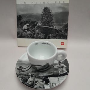 Può includere: Un set tazza e piattino da espresso bianco della collezione Illy, con un design paesaggistico in bianco e nero. La tazza ha un manico arrotondato e le parole "illy collection" stampate all'interno. Un libro intitolato "IN PRINCIPIO" è sullo sfondo.