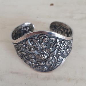 Op de afbeelding: Een zilveren manchetarmband met een sierlijk bloemen- en vogelontwerp. De armband is ongeveer 6,35 cm breed en heeft gedetailleerde gravures over het oppervlak. Het open ontwerp maakt het gemakkelijk om te dragen.