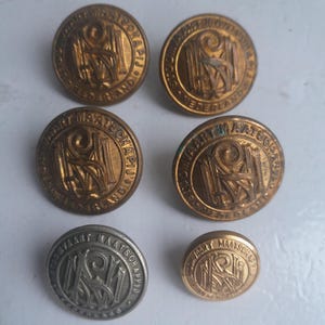 Op de afbeelding: Een verzameling vintage knopen in messing en zilverkleur. De knopen hebben een cirkelvormig ontwerp met reliëftekst en een centraal monogram. De knopen variëren in grootte en zijn waarschijnlijk afkomstig van een uniform of historisch kledingstuk.