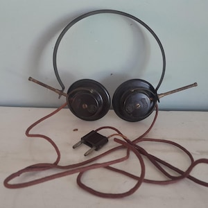 Könnte beinhalten: Vintage-Kopfhörer in Dunkelbraun mit einem dünnen Metallbügel, zwei runden Ohrmuscheln mit seitlichen Metallstäben und einem langen, ausgefransten, stoffummantelten braunen Kabel, das in einem zweipoligen Stecker für den Audioanschluss endet.