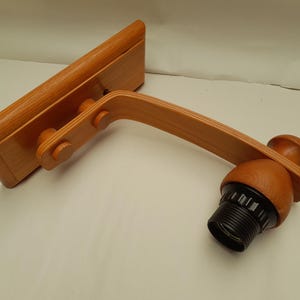 May include: Wooden wall lamp with a natural finish. It has a rectangular base, a curved arm, and a black lamp holder. The design is minimalist and functional, suitable for various interior styles. The lamp is approximately 30 cm long.