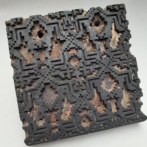Peut inclure: Un bloc de bois carré, brun foncé, avec un motif géométrique sculpté. Le bloc présente un motif pixélisé avec un motif répétitif de carrés et de lignes. Le bois a un aspect vieilli avec des zones de bois clair apparent.