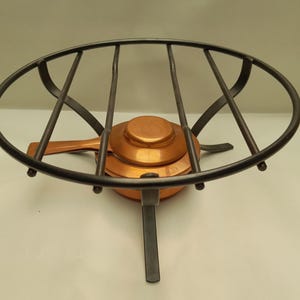 May include: A circular black metal trivet stand with four parallel crossbars and three legs, featuring a central copper-colored circular burner or heating element with a small handle extending from the side of the base component.