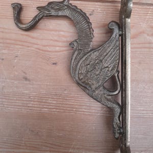 Op de afbeelding: Een wandhaak van gietijzer in de vorm van een draak met vleugels en een gekrulde staart. De haak is donkerbruin van kleur en heeft een decoratief ontwerp.