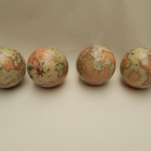 Könnte beinhalten: Vier kleine, kugelförmige Globen mit einem Vintage-Kartendesign. Die Globen haben eine Farbpalette aus Beige und Pfirsich, mit detaillierten Illustrationen von Kontinenten und Ozeanen. Sie sind in einer Reihe vor einem neutralen Hintergrund angeordnet.