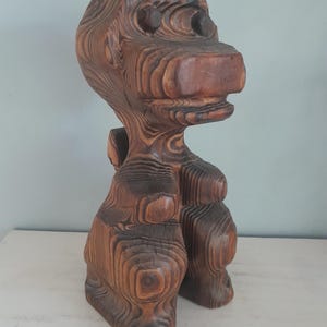 May include: A hand-carved wooden sculpture of a stylized creature with a large head and prominent snout. The figure features a dark brown finish with visible wood grain patterns throughout its body, showcasing a layered, textured appearance.