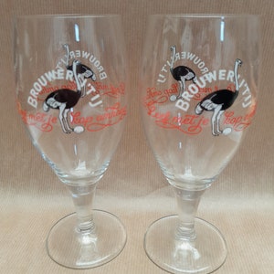 May include: Two clear stemmed beer glasses. Each glass features a cartoon ostrich illustration with the text "BROUWERIJ" and other text in red. The glasses have a wide, round base and a slender stem.
