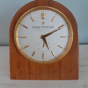 May include: A vintage Girard-Perregaux clock with a wooden base and a gold-tone bezel. The white clock face features gold-tone hands and markers, with the brand name and "Fine Watches since 1791" printed on it. The clock measures approximately 6 inches tall.