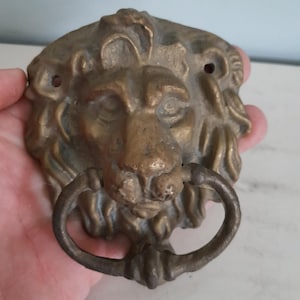 Peut inclure: Un heurtoir de porte vintage en laiton en forme de tête de lion avec une poignée en anneau. Le visage du lion est détaillé avec une crinière et des traits du visage texturés. Le heurtoir est de couleur or antique chaud.