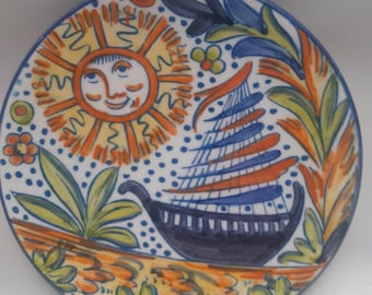 May include: A ceramic plate with a hand-painted design featuring a sun with a face, a sailboat, and floral motifs in shades of blue, orange, yellow, and green. The plate is signed "Manises".