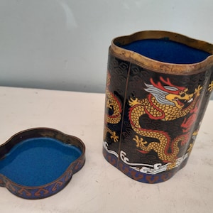 Op de afbeelding: Een zwarte en gouden cilindrische doos met een deksel. De doos is versierd met een kleurrijk drakenontwerp. Het deksel is ook versierd met een drakenontwerp. De doos is gemaakt van metaal en heeft een blauwe binnenkant.