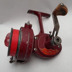 Puede incluir: Un carrete de pesca rojo vintage con un mango de madera y el texto "DAM Quick Junior". El carrete tiene una bobina roja con una banda roja y detalles plateados. El carrete está diseñado para la pesca.
