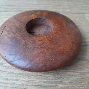 May include: A round, wooden candle holder with a central circular opening. The holder is made of a dark brown wood with visible grain and natural imperfections. The candle holder is approximately 5 inches in diameter.