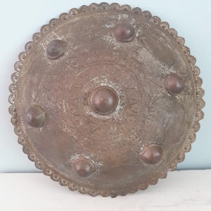 Può includere: Un vassoio circolare in metallo color rame ossidato con bordo smerlato e una cupola centrale rialzata. La superficie è decorata con cinque cupole più piccole disposte a cerchio, circondate da intricati motivi geometrici incisi.