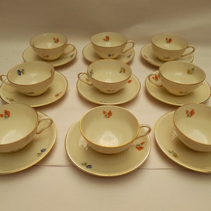 Op de afbeelding: Set van tien witte theekopjes en schoteltjes met een bloemenpatroon in tinten oranje, blauw en geel. De kopjes en schoteltjes hebben een gouden rand.