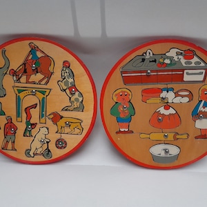 Può includere: Due puzzle circolari in legno con illustrazioni colorate. Il primo puzzle presenta un tema da circo con un clown, un elefante, un leone e un cavallo. Il secondo puzzle presenta un tema da cucina con una stufa, un lavandino e una persona che cucina.