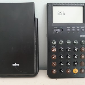 Peut inclure: Calculatrice Braun noire avec étui de protection. L'écran affiche le nombre 856. Elle possède des boutons marron et noirs, dont un bouton "start". Le logo Braun est visible.