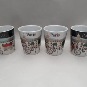 Puede incluir: Cuatro tazas de café de cerámica blanca con temática de París. Cada taza presenta un diseño de paisaje urbano con una bicicleta, señales de tráfico y la palabra "París". La parte superior de cada taza tiene una banda negra.