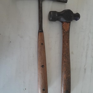 May include: Two vintage hammers with wooden handles. The hammer on the left has a curved head and the hammer on the right has a ball-peen head.