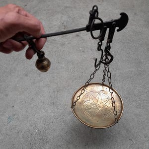 Könnte beinhalten: Eine handgehaltene Messingwaage mit einem horizontalen Balken, einem verschiebbaren Gegengewicht, einem zentralen Hakenmechanismus und einer runden Messingschale, die an drei Ketten hängt. Die Schale ist mit einem geometrischen Sternmuster graviert.