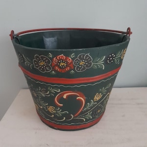 Può includere: Un secchio in legno verde scuro con un manico in metallo rosso e fasce dipinte di rosso lungo il bordo superiore e inferiore. L'esterno è decorato con motivi floreali dipinti a mano in rosso, giallo e bianco, con viticci e foglie.