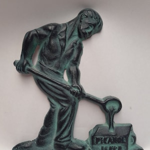 以下が含まれることがあります： 溶けた金属を型に注ぐ人の緑色の金属製フィギュア。型には「FRANK HOPE」という文字が刻まれています。