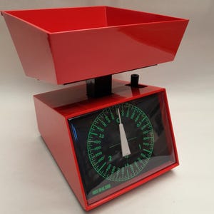 May include: A red kitchen scale with a black dial and a white needle. The dial has green markings and numbers. The scale has a red tray on top.